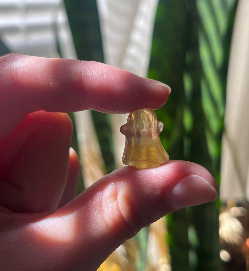 May include: A small, translucent yellow ghost figurine. The ghost has a simple, rounded shape with a small head and a flowing body. The figurine is held between two fingers, with a blurred green background.