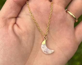 May include: A gold-colored chain necklace with a crescent moon pendant. The moon pendant is white with purple hues and has a gold-colored cap. The necklace is displayed on a hand.