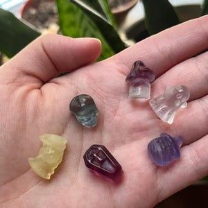 May include: A collection of small, carved crystal figurines in various shapes, including a skull, mushroom, ghost, coffin, pumpkin, and a bat-like figure. The crystals are translucent and come in shades of purple, yellow, and clear.