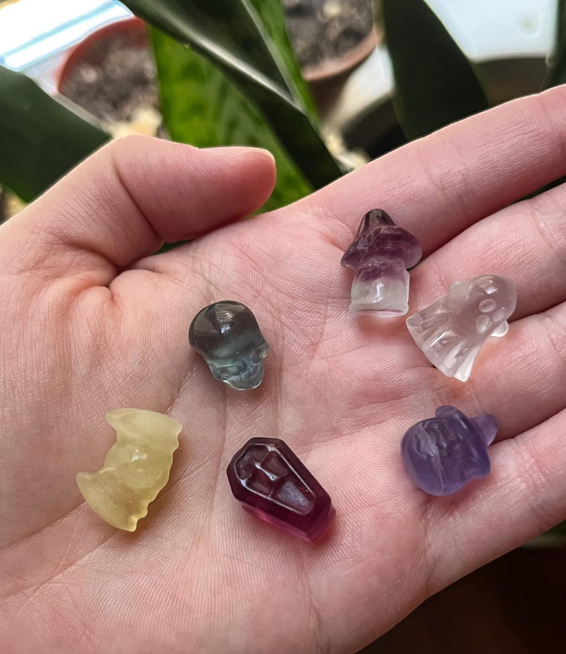 May include: A collection of small, carved crystal figurines in various shapes, including a skull, mushroom, ghost, coffin, pumpkin, and a bat-like figure. The crystals are translucent and come in shades of purple, yellow, and clear.