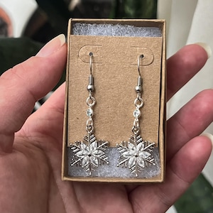 May include: Silver snowflake dangle earrings with clear crystal accents. The earrings are packaged in a brown box.