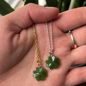 May include: Two necklaces with four-leaf clover pendants. One necklace has a gold chain, and the other has a silver chain. The clover pendants are a deep green color and appear to be made of a smooth, polished material. The necklaces are held in a hand.