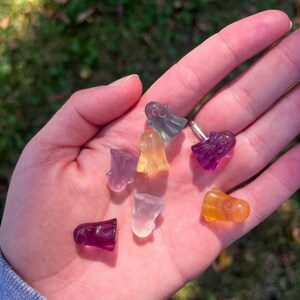 May include: A collection of small, colorful ghost-shaped crystal figurines. The crystals are in shades of purple, yellow, orange, and clear. The figurines are held in a hand against a blurred green background.