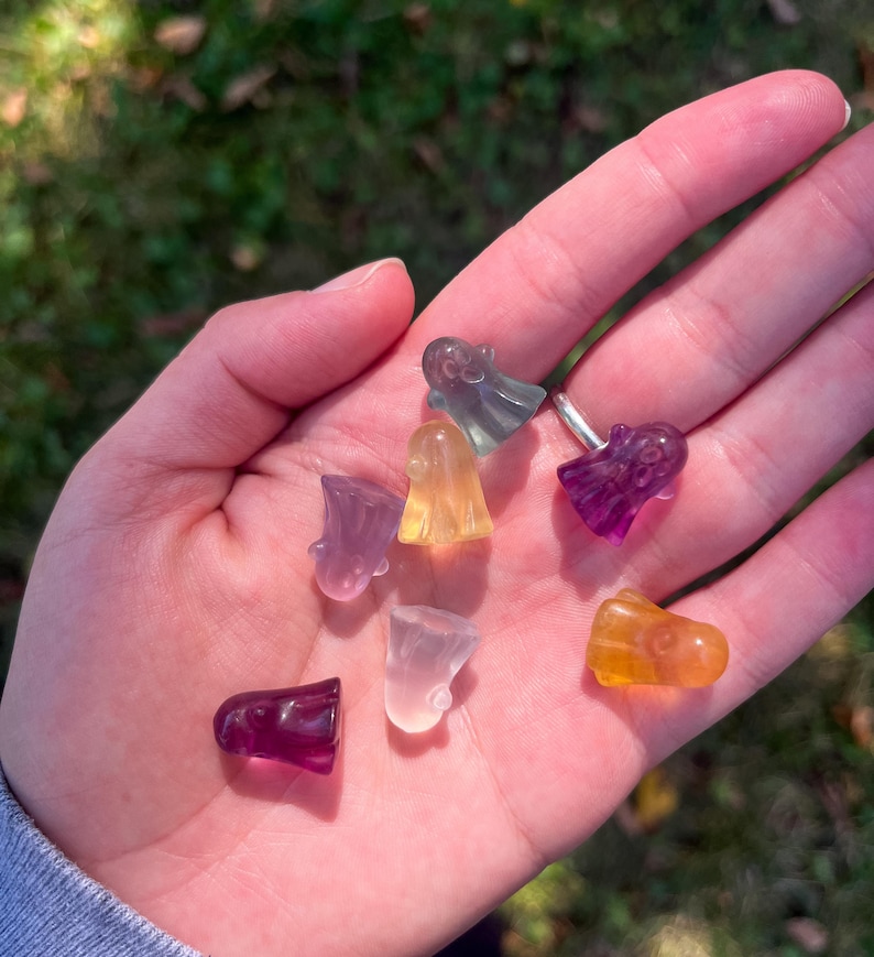 May include: A collection of small, colorful ghost-shaped crystal figurines. The crystals are in shades of purple, yellow, orange, and clear. The figurines are held in a hand against a blurred green background.