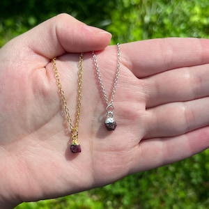 May include: Two necklaces with raw ruby pendants. One necklace has a gold chain, and the other has a silver chain. The ruby pendants are a deep red color and have a rough, natural texture. The necklaces are displayed against a blurred green background.