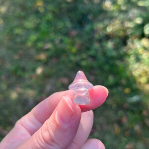 May include: A small, translucent mushroom figurine with a pinkish-purple cap and a clear stem. The mushroom is held between fingers against a blurred green background, suggesting an outdoor setting. The figurine is likely made of glass or crystal.