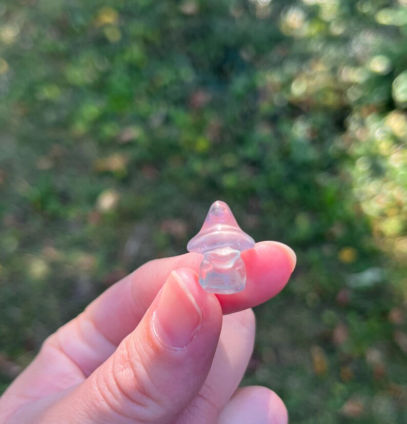 May include: A small, translucent mushroom figurine with a pinkish-purple cap and a clear stem. The mushroom is held between fingers against a blurred green background, suggesting an outdoor setting. The figurine is likely made of glass or crystal.