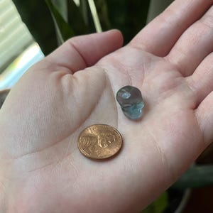 May include: A small, translucent blue skull figurine is held in a hand next to a penny. The skull is detailed with eye sockets and a nose cavity. The penny is copper-colored and shows the profile of Abraham Lincoln.
