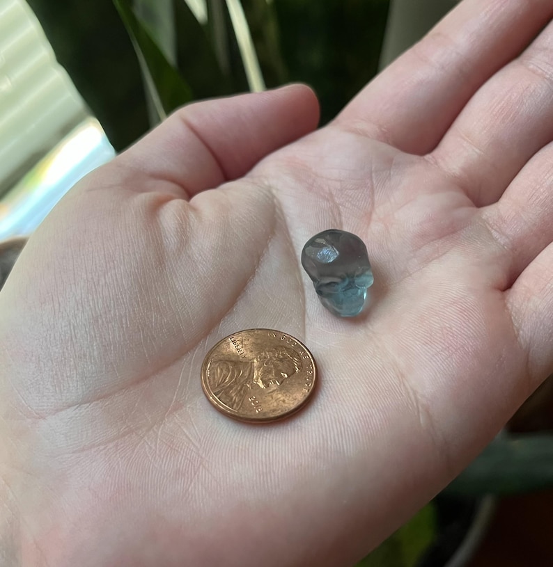 May include: A small, translucent blue skull figurine is held in a hand next to a penny. The skull is detailed with eye sockets and a nose cavity. The penny is copper-colored and shows the profile of Abraham Lincoln.