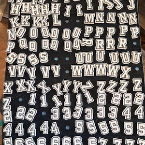 Peut inclure: Une collection de patchs en forme de lettres et de chiffres de style universitaire, blancs et noirs, disposés sur un panneau d'affichage noir. L'ensemble comprend plusieurs exemplaires de chaque lettre de A à Z et de chaque chiffre de 0 à 9.