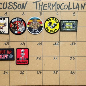Op de afbeelding: Een verzameling van zeven geborduurde strijkpatches gerangschikt op een rasterbord met het label ECUSSON THERMOCOLLANT. De patches bevatten fitness-thema afbeeldingen en teksten waaronder SOUR CLEAN, CROSSFIT, SHUT UP AND SQUAT, NO PAIN NO GAIN, BENCH PRESS, SHUT UP AND LIFT en DEADLIFT.
