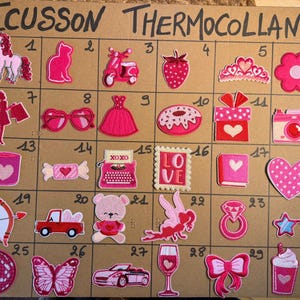 Peut inclure: Un panneau de présentation en carton intitulé ECUSSON THERMOCOLLANT présentant trente écussons brodés numérotés aux motifs roses et rouges variés, incluant une licorne, un chat, un scooter, une fraise, un diadème, une fleur, des lunettes et un cœur.