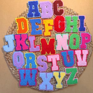 Könnte beinhalten: Eine Sammlung farbenfroher, gestickter Buchstaben in verschiedenen Farben, darunter Blau, Orange, Rosa und Grün. Jeder Buchstabe hat einen kontrastierenden Rand und ist auf einer gewebten, kreisförmigen Matte angeordnet.