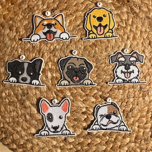 Op de afbeelding: Een verzameling van zeven geborduurde hondenpatches, elk met een ander ras met de poten rustend op een horizontale lijn. De patches zijn genummerd van één tot zeven en tonen verschillende hondenkoppen, waaronder een corgi, golden retriever en mopshond.