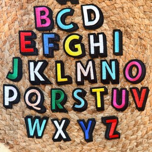 Op de afbeelding: Een complete set van zesentwintig individuele alfabetletter-patches, elk voorzien van een gedurfd, kleurrijk karakter met een dikke zwarte rand. De letters zijn in rijen gerangschikt en tonen een verscheidenheid aan levendige kleuren, waaronder rood, blauw, groen, geel, roze en wit.