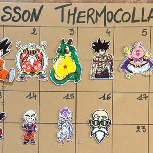 Peut inclure: Un ensemble de dix écussons thermocollants représentant divers personnages d'anime de Dragon Ball, dont Goku, Tortue Géniale, Shenron, Majin Buu, Piccolo, Krillin et Freezer, disposés sur une grille portant l'inscription ECUSSON THERMOCOLLANT.