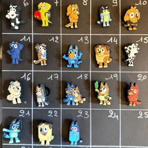 Può includere: Una collezione di ventitré ciondoli per scarpe a forma di cani dei cartoni animati, disposti in una griglia su una lavagna nera numerata da uno a trenta. Ogni ciondolo presenta un design di personaggio distinto con varie pose e colori.