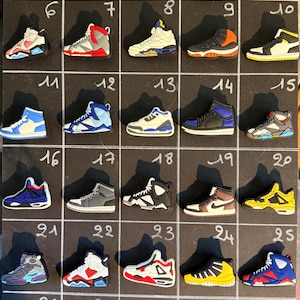 Puede incluir: Una colección de treinta dijes con forma de zapatillas deportivas dispuestos en una cuadrícula sobre un tablero negro, numerados del 1 al 30. Los dijes presentan varias combinaciones de colores que incluyen blanco, negro, azul, rojo, amarillo, naranja y gris.