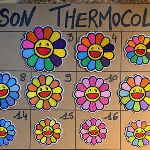 Può includere: Una collezione di undici toppe ricamate a forma di fiore con facce sorridenti, disposte su una griglia con la scritta ECUSSON THERMOCOLLANT. Le toppe presentano varie combinazioni di colori, tra cui arcobaleno, blu, rosa e giallo.