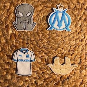 Peut inclure: Quatre écussons brodés sur une surface tissée. Les écussons comprennent un extraterrestre gris, un emblème bleu et blanc, un maillot blanc avec des accents bleus et le texte "CMA CGM", et un geste de la main beige.