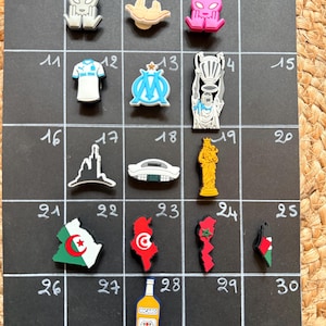 Peut inclure: Une collection de breloques décoratives, dont un extraterrestre, une voiture, un trophée et des contours de pays. Les breloques sont de différentes couleurs et motifs, disposées sur une grille noire. L'image comprend également une bouteille de Ricard.