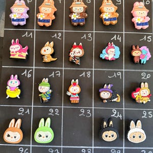Peut inclure: Une collection de trente breloques en silicone numérotées pour chaussures, représentant divers personnages de lapins de dessin animé dans différentes tenues, couleurs et poses, disposées en grille sur un fond noir.