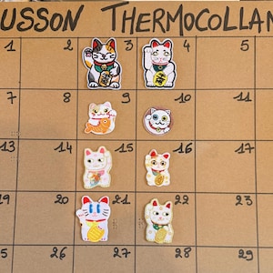 Peut inclure: Huit écussons thermocollants brodés représentant divers chats porte-bonheur Maneki-neko dans des tons blanc, noir, orange et doré. Les écussons sont disposés sur un quadrillage marron avec le texte ECUSSON THERMOCOLLANT en haut.