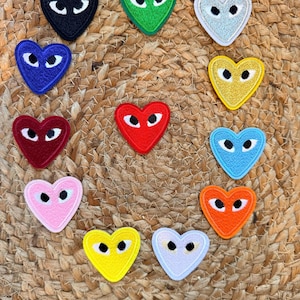 Op de afbeelding: Een verzameling van twaalf hartvormige geborduurde patches in verschillende kleuren, waaronder zwart, groen, zilver, blauw, geel, rood, lichtblauw, roze, oranje en lichtpaars. Elk hart is voorzien van een paar witte ogen met zwarte pupillen.
