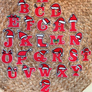 Peut inclure: Une collection de vingt-six écussons brodés rouges représentant l'alphabet, chacun orné d'accents festifs tels que des bonnets de Père Noël, des bois de renne, des nœuds et des chaussettes. Les lettres sont disposées de A à Z sur une surface tressée.