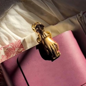 Peut inclure: Un clip décoratif doré en forme de main, possiblement un signet ou une attache, repose sur une surface en cuir violet. Le design du clip comprend des détails complexes, sur un fond de tissu blanc et à motifs.