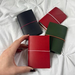 Op de afbeelding: Vier kleine, leren notitieboekjes in zwart, rood en groen, elk vastgezet met een elastiek. De notitieboekjes zijn gerangschikt op een wit, getextureerd oppervlak. Een rood notitieboekje wordt door een hand vastgehouden.