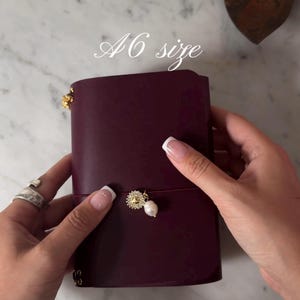 Pode incluir: Um diário A6 bordô com fecho dourado e um pingente de pérola. O diário está aberto, mostrando seu interior. O texto "A6 size" está escrito em itálico na parte superior da imagem.