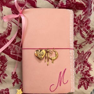 Puede incluir: Un cuaderno rosa claro con un borde de encaje blanco en el lado derecho y una cinta rosa atada arriba a la izquierda. La cubierta presenta una letra M bordada en rosa y una banda elástica rosa sostiene tres dijes dorados: un corazón, un lazo y una luna.