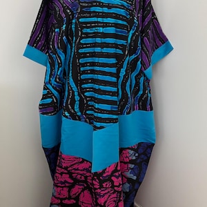 Op de afbeelding: Een levendige, patroon caftan jurk op een mannequin. De jurk heeft een V-hals en korte mouwen. De stof toont een mix van zwarte, blauwe, paarse en roze abstracte ontwerpen. De jurk is een los, vloeiend kledingstuk.