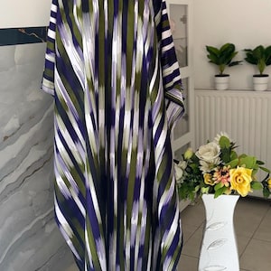 Op de afbeelding: Een lange, vloeiende jurk met een verticaal streepjespatroon in wit, olijfgroen en paars. De jurk wordt op een mannequin gepresenteerd. De jurk heeft korte mouwen en een brede halslijn. De achtergrond omvat een vaas met bloemen.