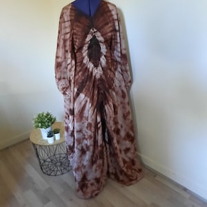 Op de afbeelding: Een lange, vloeiende tie-dye jurk in bruin- en wittinten. De jurk heeft een V-hals en lange mouwen. De stof heeft een zachte, lichtgewicht uitstraling, met een patroon dat vanuit het midden straalt.