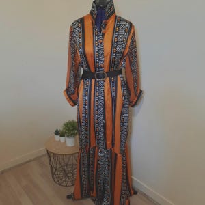 Op de afbeelding: Een lange oranje jurk met een zwart-wit geometrisch patroon, lange mouwen en een hoge kraag. De jurk is in de taille vastgemaakt met een zwarte riem en heeft een geplooid zoom. De jurk wordt op een mannequin getoond.