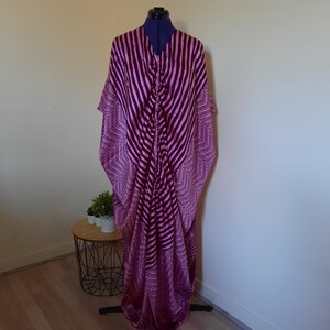 Op de afbeelding: Een lange, magenta en paars gekleurde kaftan jurk. De jurk heeft een V-hals en een verticaal gestreept ontwerp dat overgaat in een geometrisch patroon aan de zoom en mouwen. Het kledingstuk wordt getoond op een blauwe paspop.