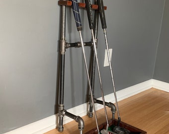 May include: A black metal golf club rack with three golf clubs and a wooden base with a green felt insert for storing golf putters. The rack is mounted to a wall.