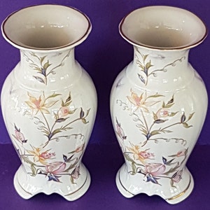 May include: A pair of white ceramic vases featuring floral patterns in pink, yellow, and green. Each vase has a flared top with a brown rim and a decorative base with gold trim. The floral design includes stylized flowers and leaves.