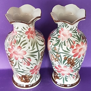 May include: Two white ceramic vases with a floral design. The vases have a ruffled rim and are decorated with pink flowers, green leaves, and brown accents. The vases are approximately 25 cm tall.