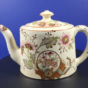 May include: Cream ceramic teapot with lid, handle, and spout. Decorated with pink and yellow floral patterns and brown accents. The design includes a circular floral motif and a geometric border. A classic tea accessory.