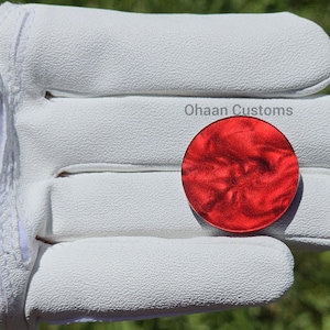 May include: A red, round, marbled, resin blank for a guitar pick. The blank is being held in a white leather glove. The text "Ohaan Customs" is visible in the image.