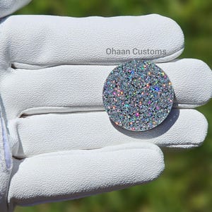 Peut inclure: Une pièce ronde, argentée, scintillante, irisée et décorative avec une surface texturée. La pièce est recouverte de petits strass chatoyants et colorés. "Ohaan Customs" est écrit en noir sur le fond.