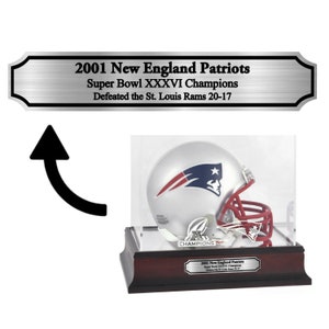 May include: A silver football helmet with a red facemask and a blue and white logo is displayed in a clear acrylic case on a wooden base. The base has a silver plaque that reads "2001 New England Patriots Super Bowl XXXVI Champions Defeated the St. Louis Rams 20-17".