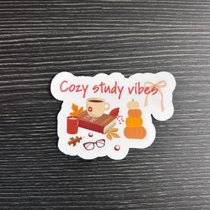 May include: A die-cut sticker featuring the text "Cozy study vibes" in red script. The illustration includes a tan mug on a book, a red candle, black-rimmed glasses, autumn leaves, a stack of three orange pumpkins, and a light tan ribbon.