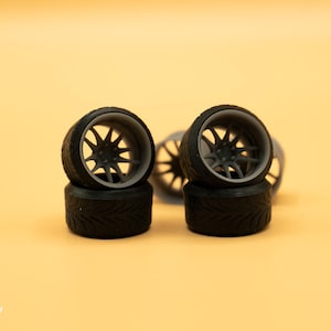 May include: Close-up of four black and gray model car wheels with a detailed spoke design. The wheels are arranged in two stacks of two, with the front wheels facing the camera.