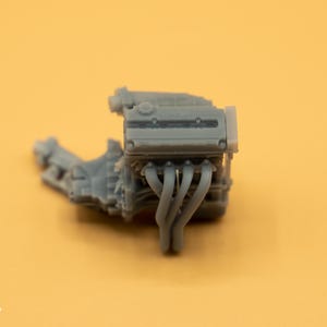 Puede incluir: Un modelo de motor de coche de plástico gris con un colector de admisión y tubos de escape detallados.