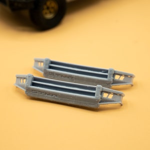 May include: Two grey model car bumpers, each with the text "STMA". The bumpers are rectangular with three horizontal bars, designed for a miniature vehicle. The background is a solid yellow colour.