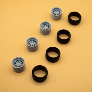 Puede incluir: Un conjunto de seis ruedas de coche en miniatura, cuatro neumáticos negros y dos ruedas de color azul claro con un diseño de radios.